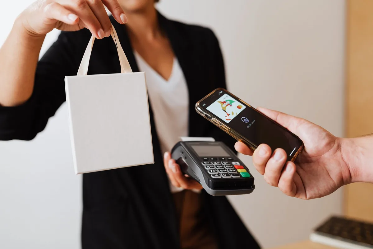 Customer paying with a smartphone at a card terminal while receiving a shopping bag, representing payment and fintech apps