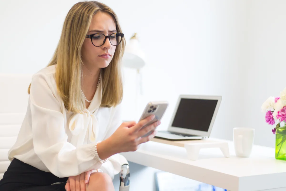 Woman at a desk checking a smartphone next to a laptop